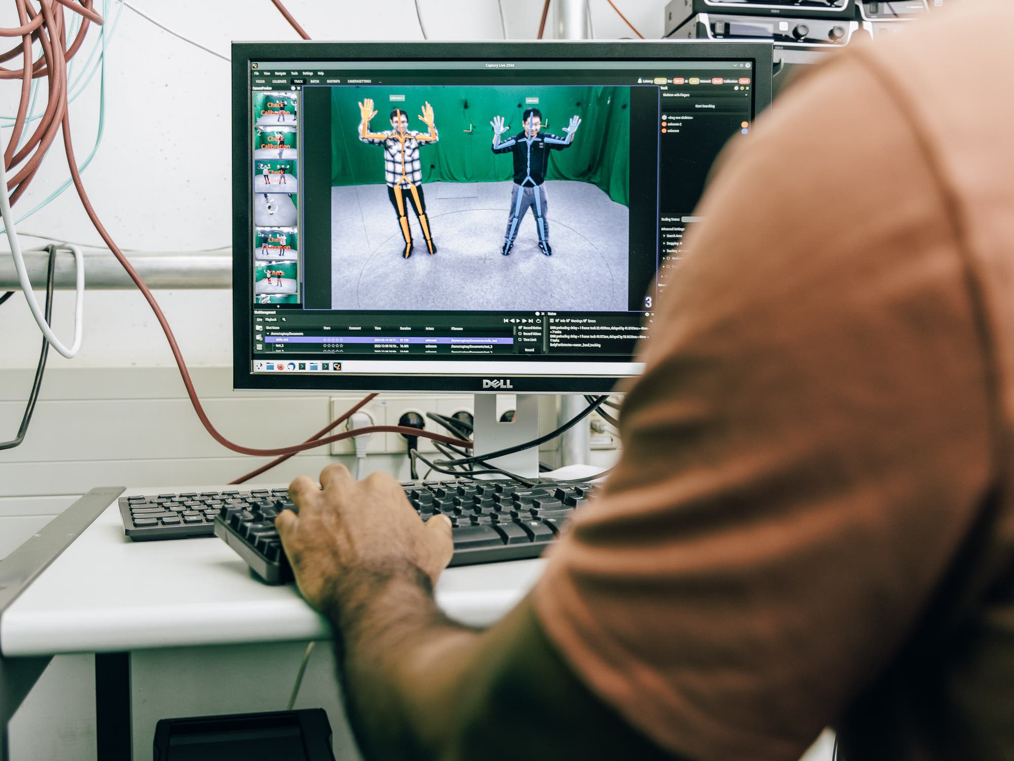 Eine Person sitzt am Computer und filmt zwei Menschen in einem grünen Raum: Die zwei Menschen sind mit Linien überzogen, die rudimentär wie Strichmännchen ihre Figuren darstellen.