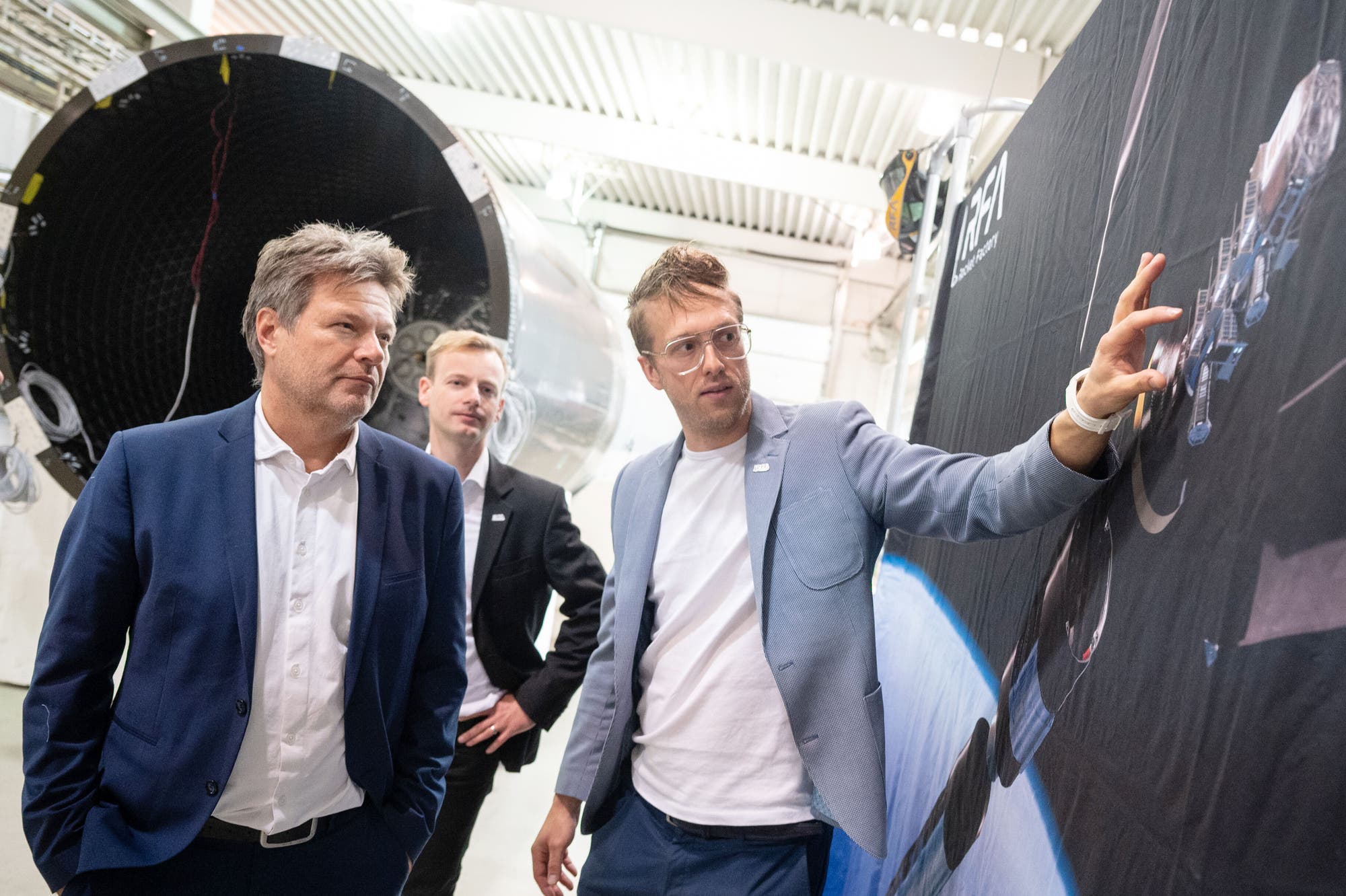 Drei Männer in Anzügen stehen in einer industriellen Umgebung. Einer von ihnen zeigt auf ein großes Bild, das Raumfahrttechnologie darstellt. Im Hintergrund ist ein Teil eines großen zylindrischen Objekts zu sehen, das wie ein Raketenteil aussieht. Die Szene vermittelt eine Diskussion über Raumfahrt oder Technologie.