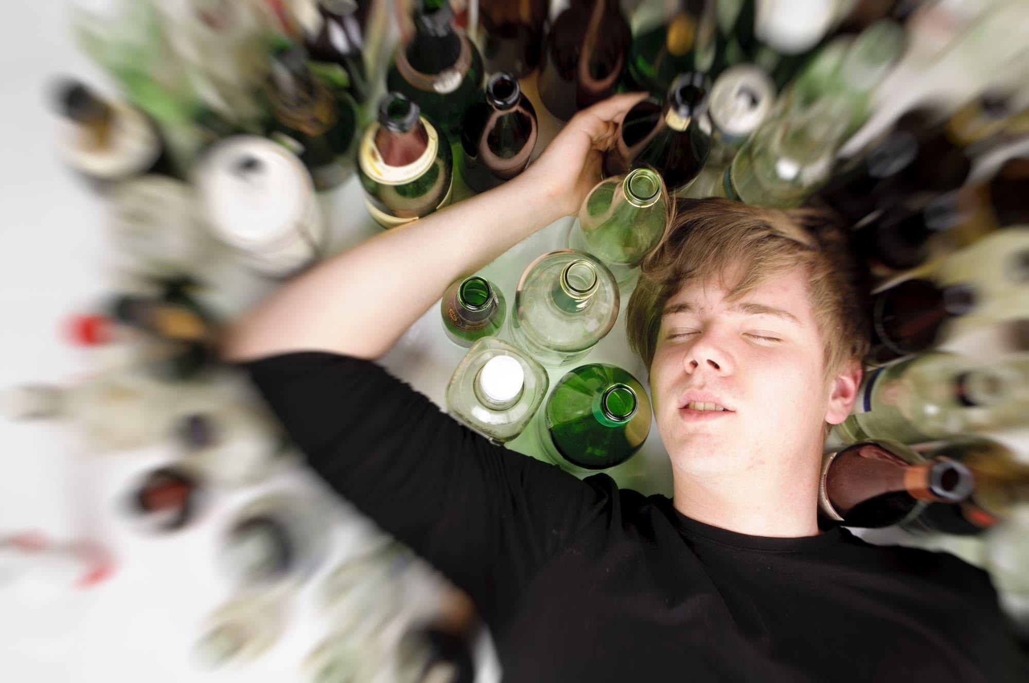 Ein männlicher Jugendlicher liegt im Alkoholrausch schlafend neben vielen leeren Flaschen auf dem Boden.