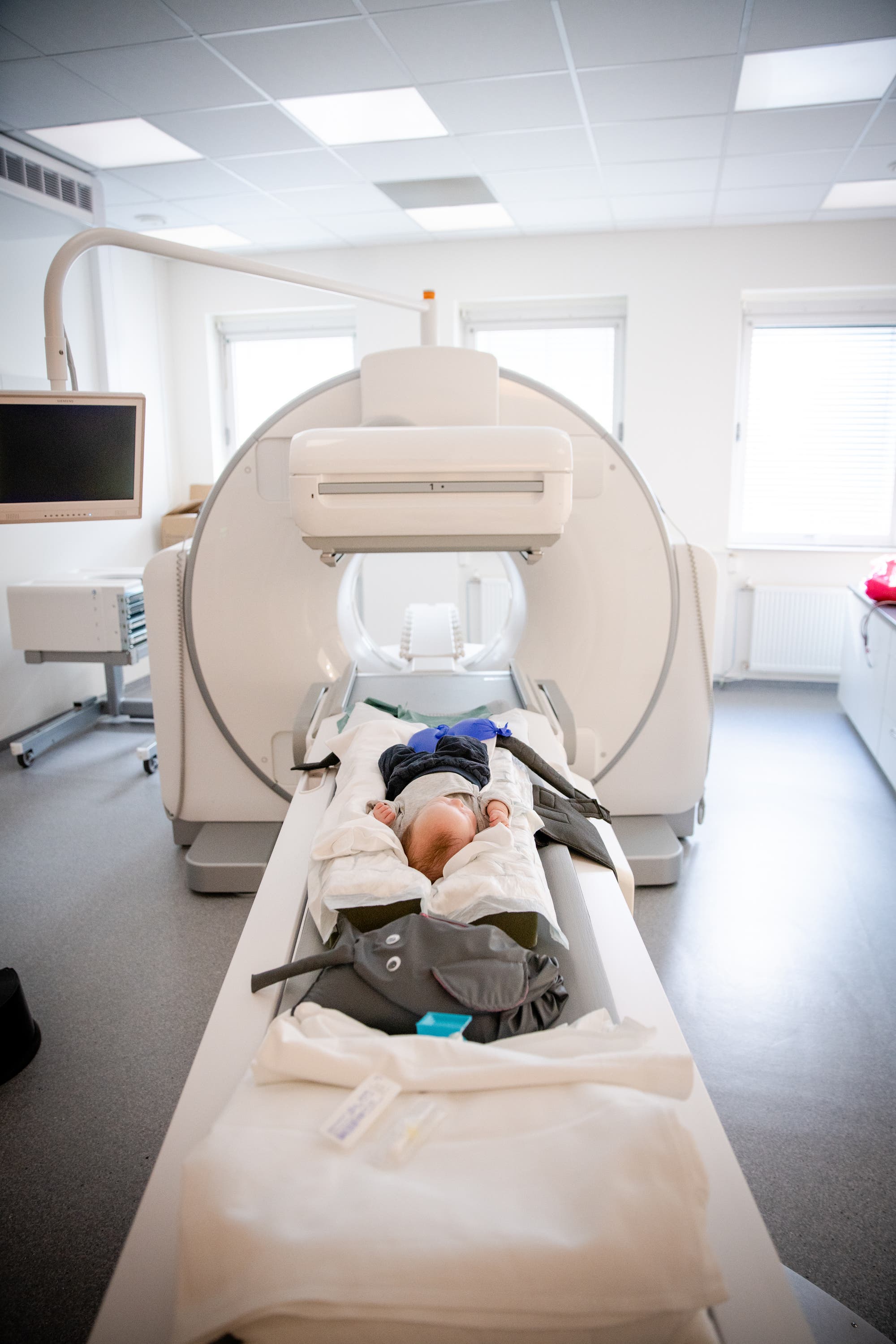Ein Baby liegt auf dem Rücken auf einem Untersuchungstisch, der in einen großen medizinischen Scanner geschoben wird. Der Raum ist hell erleuchtet und modern eingerichtet. Das Baby trägt eine Schutzweste. Der Scanner ist ein großes, rundes Gerät, das für bildgebende Verfahren wie MRT oder CT verwendet wird. Im Hintergrund sind Fenster und ein Monitor zu sehen.