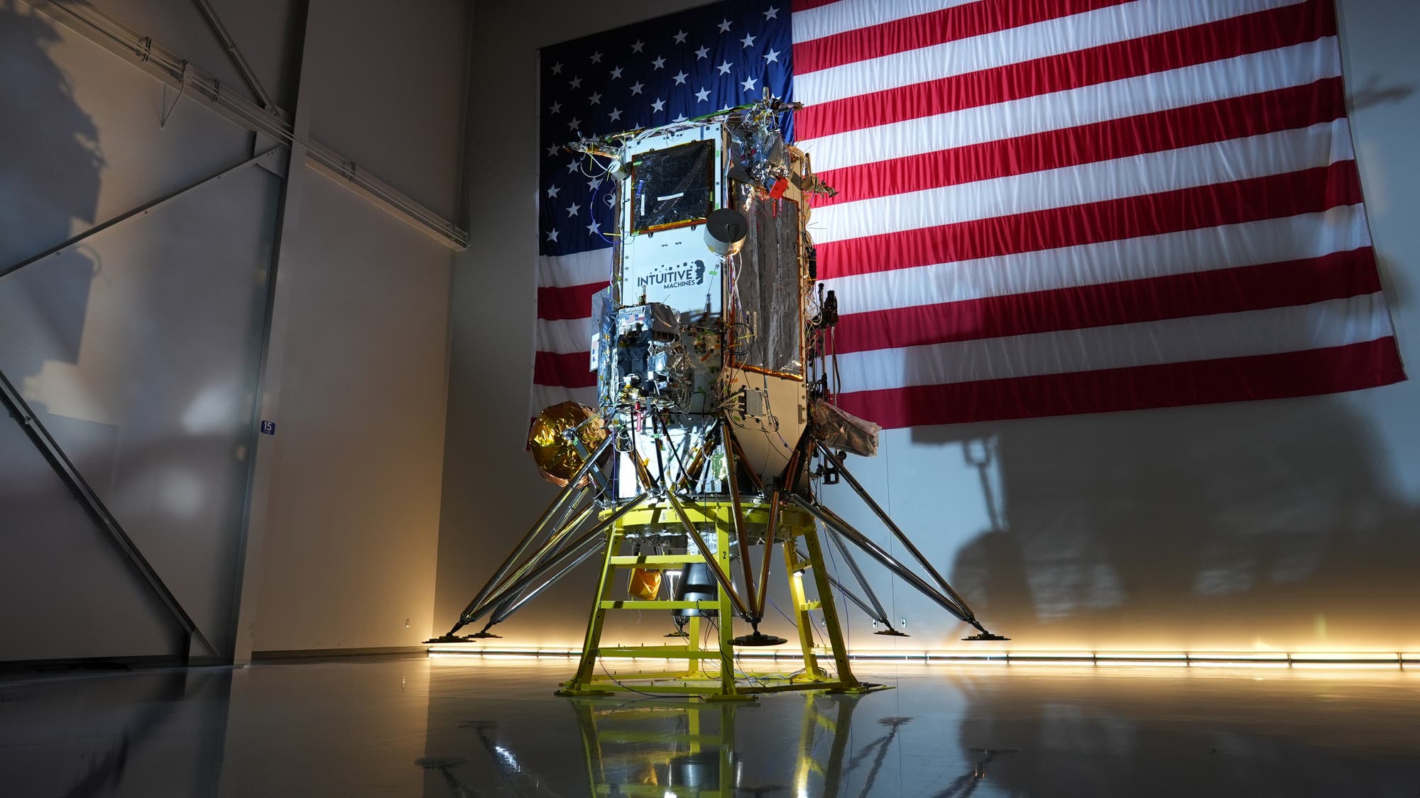 Eine Mondlandefähre steht in einem Raum vor einer großen US-Flagge. Die Landefähre ist mit verschiedenen technischen Komponenten ausgestattet und steht auf einem gelben Gestell. Auf der Seite der Landefähre ist das Logo von "Intuitive Machines" zu sehen. Der Raum ist gut beleuchtet, und die glänzende Oberfläche des Bodens spiegelt die Landefähre wider.