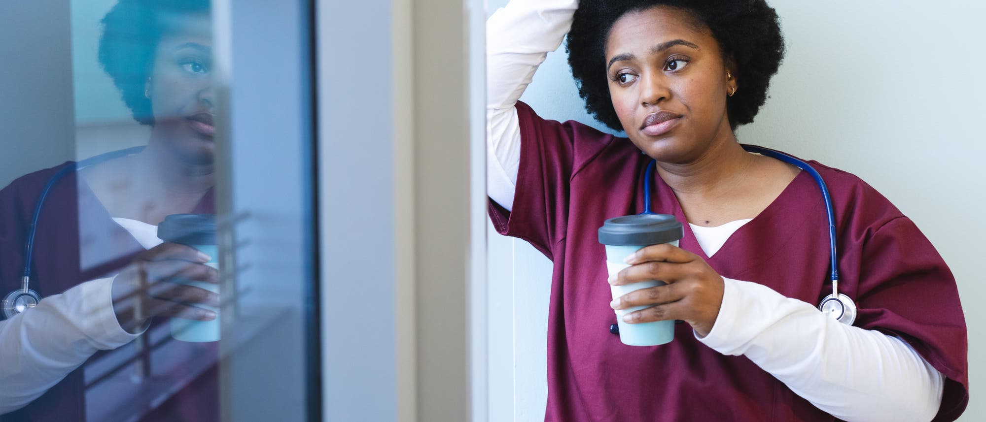 Eine Person in medizinischer Kleidung lehnt sich nachdenklich an eine Wand und hält einen Kaffeebecher. Sie trägt ein Stethoskop um den Hals. Die Reflexion der Person ist im Fenster zu sehen. Die Szene vermittelt einen Moment der Ruhe oder Reflexion in einem hektischen Arbeitsumfeld. Eine Person in medizinischer Kleidung lehnt sich nachdenklich an eine Wand und hält einen Kaffeebecher. Sie trägt ein Stethoskop um den Hals. Die Reflexion der Person ist im Fenster zu sehen. Die Szene vermittelt einen Moment der Ruhe oder Reflexion in einem hektischen Arbeitsumfeld.
