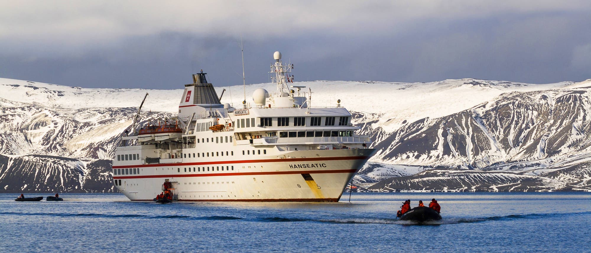 Ein großes Kreuzfahrtschiff mit dem Namen "Hanseatic" fährt durch eine ruhige, eisige Gewässerlandschaft, umgeben von schneebedeckten Bergen. Im Vordergrund bewegt sich ein kleines Schlauchboot mit mehreren Personen in Richtung des Schiffes. Der Himmel ist bewölkt, was der Szene eine kühle, arktische Atmosphäre verleiht. Ein großes Kreuzfahrtschiff mit dem Namen "Hanseatic" fährt durch eine ruhige, eisige Gewässerlandschaft, umgeben von schneebedeckten Bergen. Im Vordergrund bewegt sich ein kleines Schlauchboot mit mehreren Personen in Richtung des Schiffes. Der Himmel ist bewölkt, was der Szene eine kühle, arktische Atmosphäre verleiht.