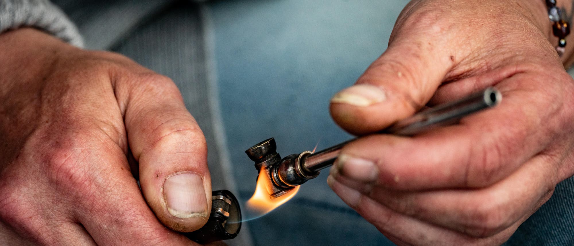 Eine Person hält eine kleine Metallpfeife in der rechten Hand und zündet sie mit einem Feuerzeug in der linken Hand an. Die Hände sind nah aufgenommen, und die Person trägt ein Armband mit bunten Perlen. Die Szene vermittelt den Akt des Rauchens.