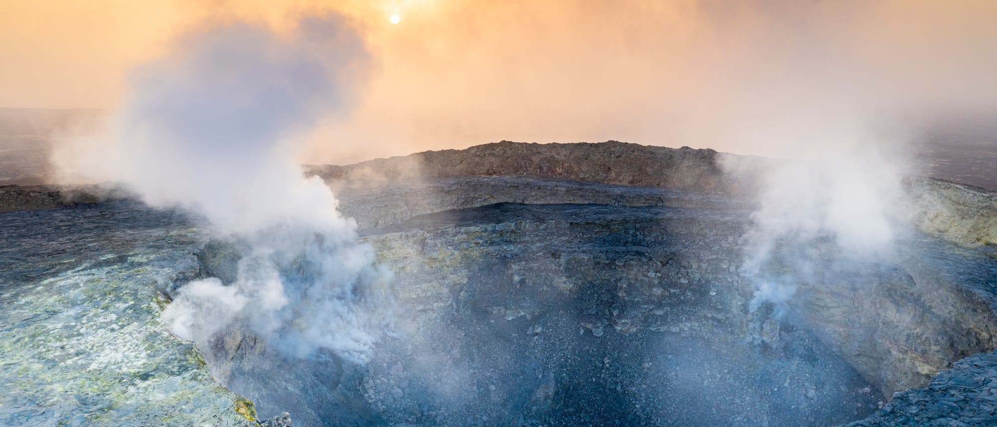 Ein aktiver Vulkan mit einem rauchenden Krater, aus dem weiße Dampfwolken aufsteigen. Der Himmel ist in warmen Gelb- und Orangetönen gefärbt, während die Sonne durch den Dunst scheint. Die vulkanische Landschaft ist von erstarrter Lava umgeben. Ein aktiver Vulkan mit einem rauchenden Krater, aus dem weiße Dampfwolken aufsteigen. Der Himmel ist in warmen Gelb- und Orangetönen gefärbt, während die Sonne durch den Dunst scheint. Die vulkanische Landschaft ist von erstarrter Lava umgeben.