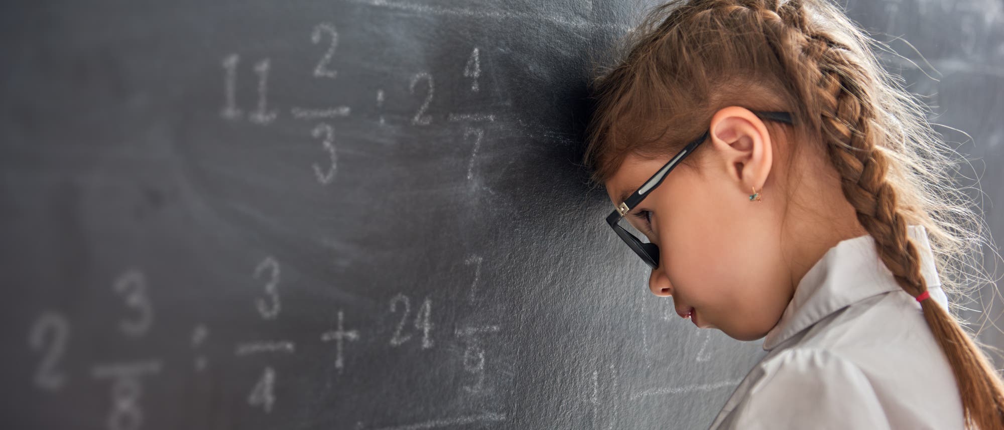 Ein Mädchen mit Brille und geflochtenem Haar lehnt seinen Kopf gegen eine Tafel, auf der mathematische Brüche und Gleichungen geschrieben sind. Es trägt eine weiße Bluse und wirkt nachdenklich oder frustriert. Ein Mädchen mit Brille und geflochtenem Haar lehnt seinen Kopf gegen eine Tafel, auf der mathematische Brüche und Gleichungen geschrieben sind. Es trägt eine weiße Bluse und wirkt nachdenklich oder frustriert.