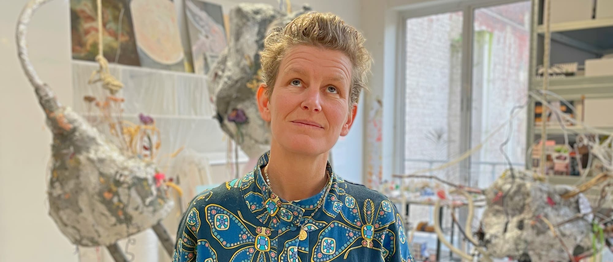 Eine Person mit kurzem Haar steht in einem Kunststudio und blickt nachdenklich nach oben. Sie trägt ein Hemd mit buntem Muster. Im Hintergrund sind Skulpturen und Kunstwerke zu sehen, die aus verschiedenen Materialien bestehen. Durch ein großes Fenster fällt Tageslicht in den Raum. Eine Person mit kurzem Haar steht in einem Kunststudio und blickt nachdenklich nach oben. Sie trägt ein Hemd mit buntem Muster. Im Hintergrund sind Skulpturen und Kunstwerke zu sehen, die aus verschiedenen Materialien bestehen. Durch ein großes Fenster fällt Tageslicht in den Raum.