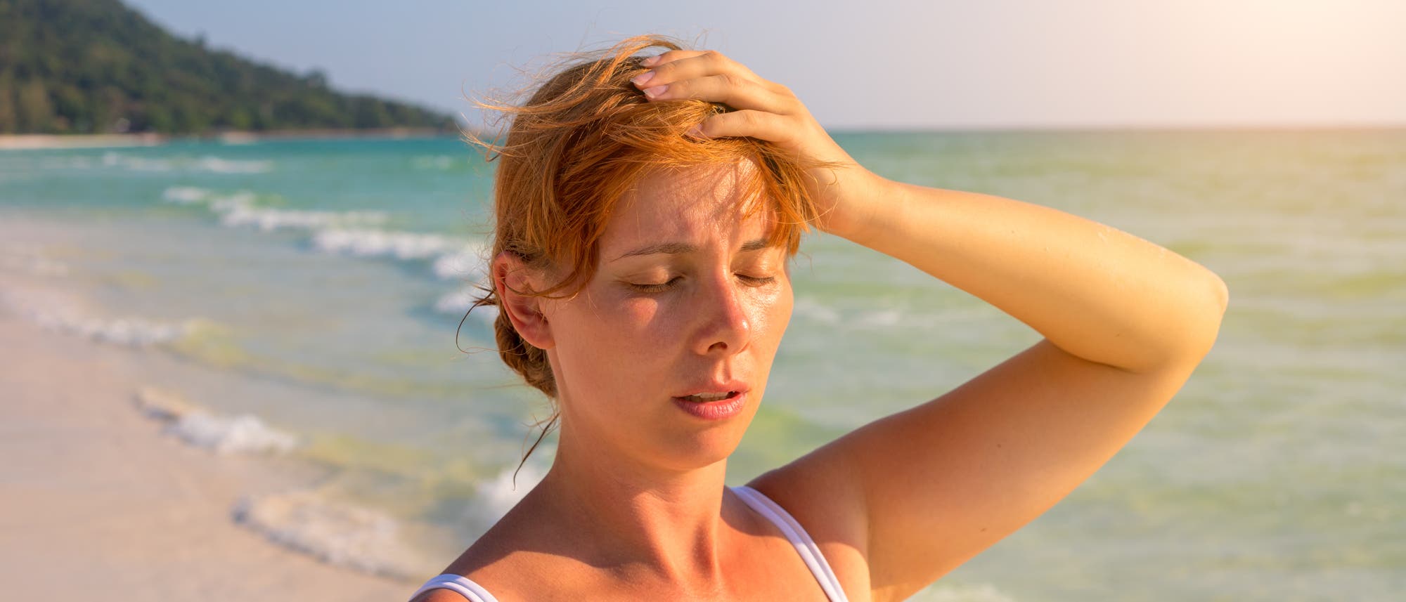 Frau steht an einem Strand in der Sonne, sieht erschöpft aus und fasst sich an den Kopf. Frau steht an einem Strand in der Sonne, sieht erschöpft aus und fasst sich an den Kopf.