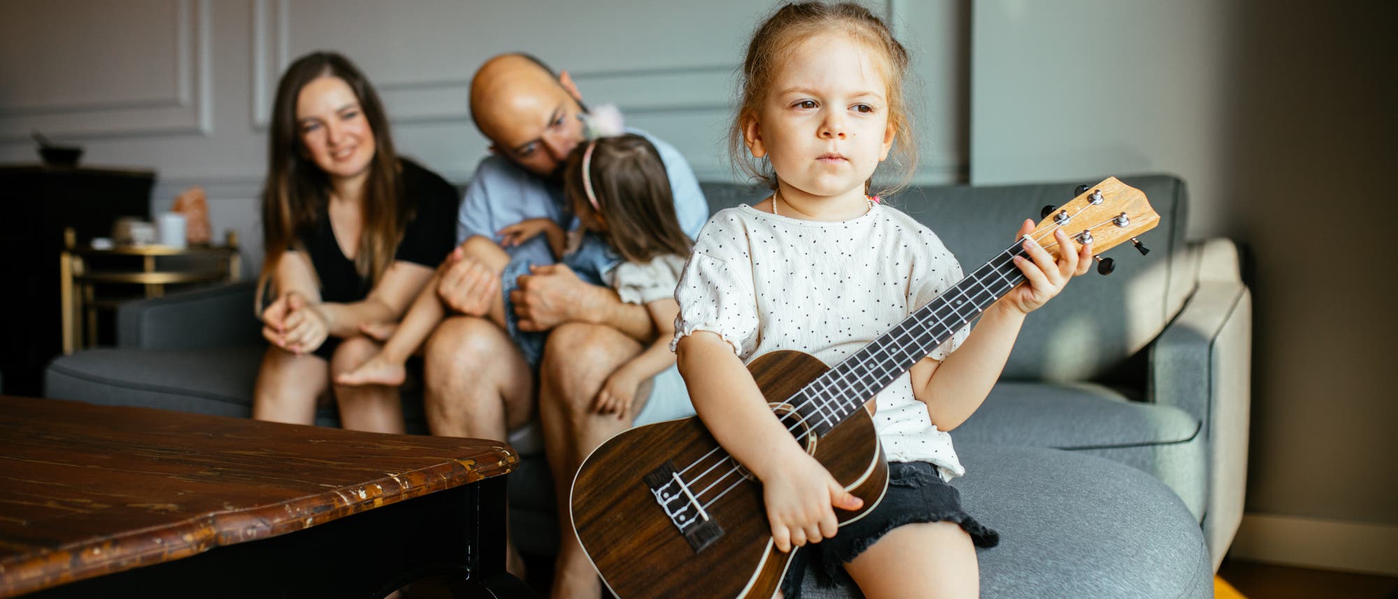 Eine Familie mit zwei Kindern sitzt im Wohnzimmer. Mutter und Vater kümmern sich um das eine Kind, während das andere Kind mit einer Gitarre auf dem Schoß traurig etwas abseits sitzt. Eine Familie mit zwei Kindern sitzt im Wohnzimmer. Mutter und Vater kümmern sich um das eine Kind, während das andere Kind mit einer Gitarre auf dem Schoß traurig etwas abseits sitzt.