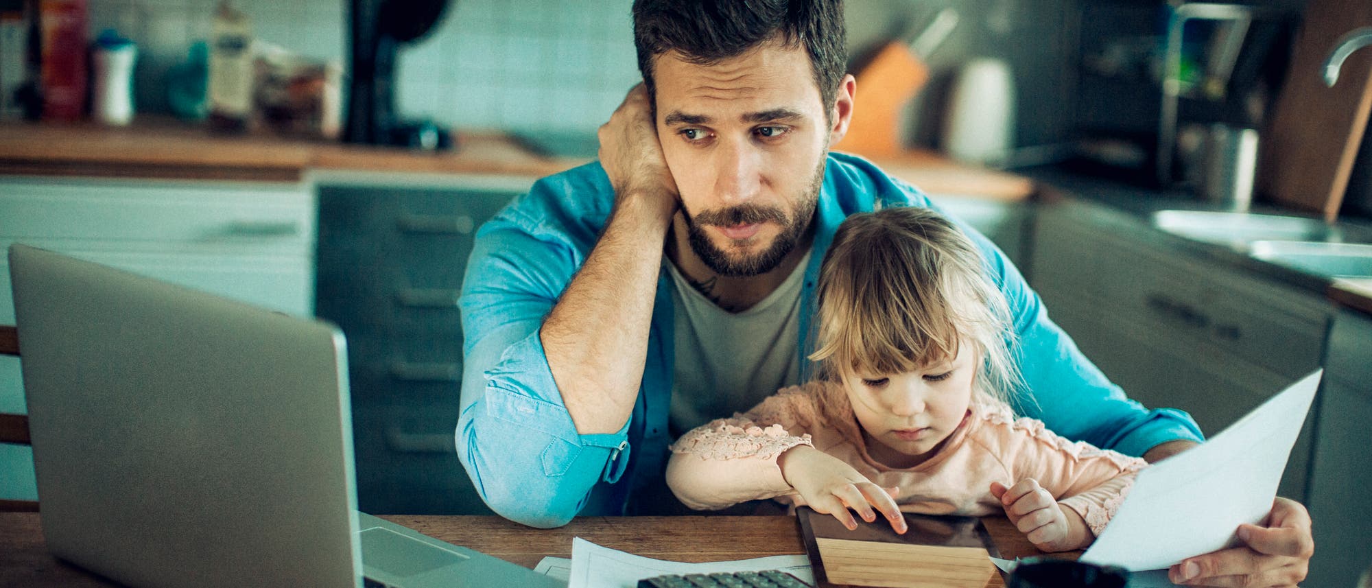 Junger Vater sitzt mit Tochter am Küchentisch und versucht dabei zu arbeiten.