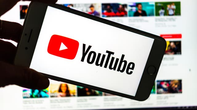 Ein Smartphone wird in der Hand gehalten und zeigt das YouTube-Logo auf dem Bildschirm. Im Hintergrund ist ein unscharfer Computerbildschirm zu sehen, der die YouTube-Webseite mit mehreren Video-Thumbnails anzeigt. Das Bild vermittelt die Nutzung von YouTube sowohl auf mobilen Geräten als auch auf Desktop-Computern.
