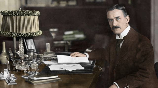 Ein Mann im Anzug sitzt an einem Schreibtisch, umgeben von Büchern und Papieren. Er hält ein Schriftstück in der rechten Hand und schaut nach links direkt in die Kamera. Auf dem Schreibtisch stehen eine verzierte Lampe, Schreibutensilien und ein Tintenfass. Im Hintergrund sind Bücherregale zu sehen. Die Atmosphäre ist ernst und erhaben.