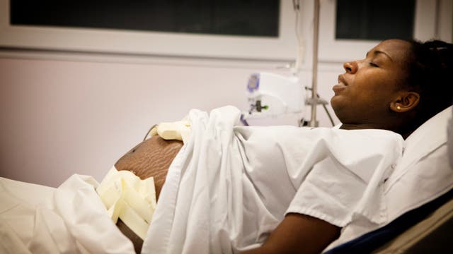 Eine schwangere Frau liegt in einem Krankenhausbett, teilweise zugedeckt mit einem weißen Laken. Sie trägt ein Krankenhaushemd und scheint sich in einer Entbindungsstation zu befinden. Neben ihr ist medizinisches Equipment sichtbar. Die Frau wirkt konzentriert und ruhig.