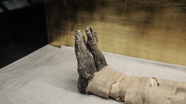 Die Aufnahme zeigt die Füße einer mumifizierten Person, die in Leinenbinden gewickelt sind. Die Mumie liegt auf einem hellen Tuch vor einem goldfarbenen Hintergrund. Die Szene vermittelt einen Eindruck von antiker Bestattungskultur.