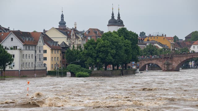Die braunen Fluten des Neckars schießen durch Heidelberg und überfluten Teile der Altstadt. Der Blick geht von Osten in Richtung alte Brücke, auf der zahlreiche menschen stehen. Links ist die Altstadt mit verschiedenen Gebäuden zu sehen, darunter auch der Turm der Heiliggeistkirche