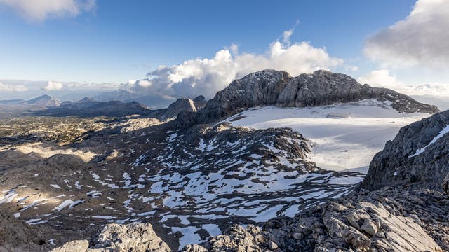 Blick auf eine weite, beeindruckende Berglandschaft in den österreichischen Alpen mit schneebedeckten Gipfeln und Felsen unter klarem blauen Himmel. Im Hintergrund sind Wolken zu sehen. Blick vom Kleinen Gjadstein in Richtung Osten, rechts im Bild ist der Schladminger Gletscher zu sehen.