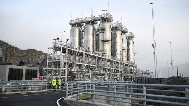 Eine industrielle Anlage mit mehreren großen silbernen Tanks und Rohrleitungen, umgeben von einem Zaun. Zwei Personen in gelben Sicherheitsjacken stehen auf einer Straße vor der Anlage und scheinen sich zu unterhalten. Im Hintergrund sind felsige Hügel zu sehen. Die Szene vermittelt den Eindruck einer modernen Industrieumgebung.