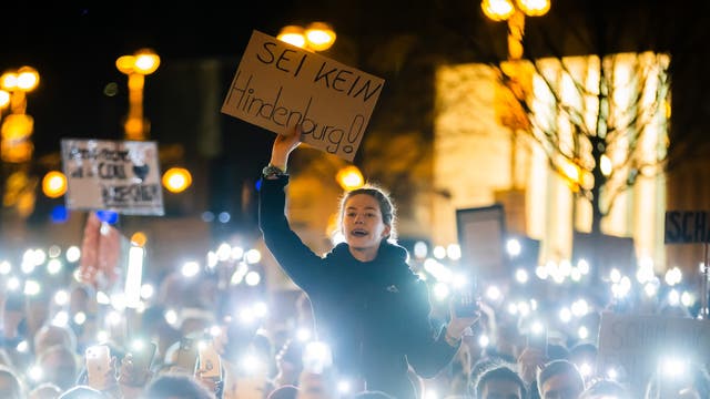 Eine junge Frau steht inmitten einer Menschenmenge bei Nacht und hält ein Schild mit der Aufschrift "Sei kein Hindenburg!" hoch. Die umstehenden Personen leuchten mit ihren Handys, wodurch ein Meer aus Licht entsteht. Im Hintergrund sind beleuchtete Straßenlaternen und unscharfe Gebäude zu sehen. Die Szene vermittelt den Eindruck einer friedlichen, aber emotional aufgeladenen Demonstration oder eines Protests.