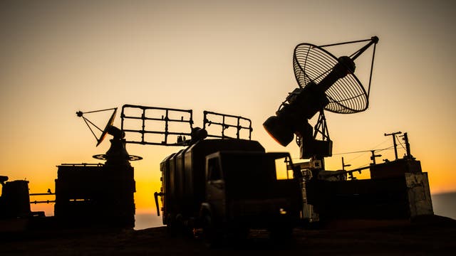 Silhouette einer militärischen Radaranlage bei Sonnenuntergang. Im Vordergrund ist ein Lastwagen zu sehen, umgeben von mehreren großen Radarantennen. Die Antennen sind auf Plattformen montiert und zeigen in verschiedene Richtungen. Der Himmel im Hintergrund ist in Gelb- und Orangetönen gefärbt.