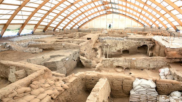 Eine archäologische Ausgrabungsstätte unter einem großen, gewölbten Schutzdach. Die frei gelegten Mauern zeigen die Struktur einer neolithischen Siedlung. Sand- und Erdsäcke sind um die Ausgrabungsbereiche herum platziert, um die Stabilität zu gewährleisten. Eine Person steht im Hintergrund und betrachtet die Ausgrabungen.