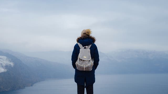 Eine Person in einem dunklen Anorak mit Kapuze und einer hellbraunen Mütze steht mit dem Rücken zur Kamera und blickt von einem Berg auf eine winterliche Landschaft mit einem See und bewölktem Himmel. Die Person hat die Hände in den Taschen und trägt einen grauen Rucksack.