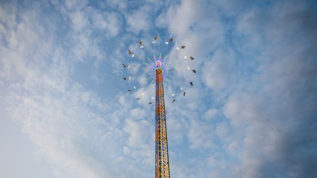 Ein hoher Kettenkarussell-Turm erstreckt sich in den Himmel, umgeben von schwebenden Sitzen, die Menschen tragen. Der Turm ist mit bunten Lichtern geschmückt, die im Kontrast zum blauen, leicht bewölkten Himmel stehen. Die Menschen scheinen in der Luft zu schweben, während sie die Fahrt genießen.