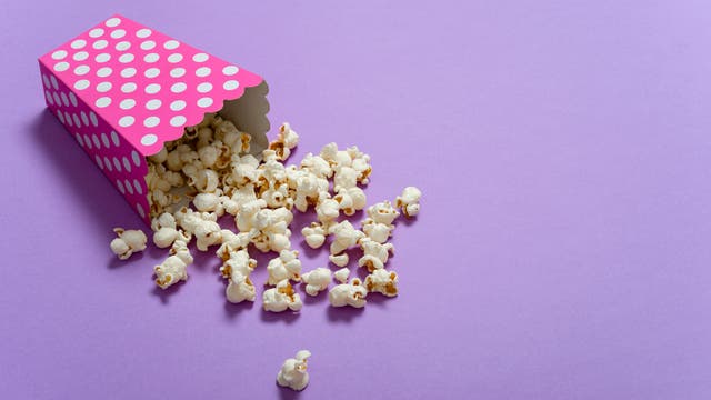Eine pinkfarbene Popcorn-Schachtel mit weißen Punkten liegt auf einer lila Oberfläche. Popcorn ist aus der Schachtel herausgefallen und verteilt sich auf dem Untergrund.