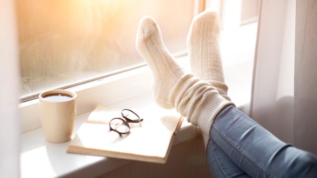 Fensterbank mit Buch und Brille darauf, einer Tasse Kaffee und hochgelegten Füßen, durchs Fenster scheint die Sonne