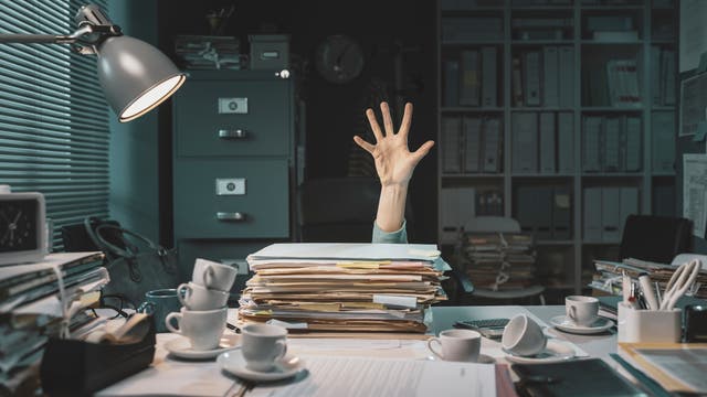 Hand schaut hinter einem Schreibtisch hervor, auf dem zahlreiche Akten liegen