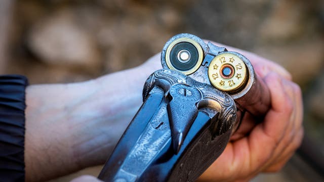 Eine Person hält eine doppelläufige Schrotflinte, die geöffnet ist, um zwei Patronen zu zeigen. Die Patronen sind mit "12" und Sternen markiert. Die Waffe hat ein kunstvolles Gravurmuster. Der Hintergrund ist unscharf und zeigt eine natürliche Umgebung.