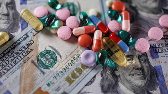Verschiedene bunte Pillen und Kapseln liegen verstreut auf mehreren 100-Dollar-Scheinen. Die Szene symbolisiert die Verbindung zwischen Geld und der Pharmaindustrie. Im Hintergrund ist das Porträt von Benjamin Franklin auf den Geldscheinen zu sehen.