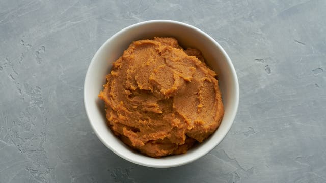Eine weiße Schüssel mit einer Portion brauner Miso-Paste, auf einer grauen, strukturierten Oberfläche platziert. Die Paste hat eine glatte, cremige Konsistenz.