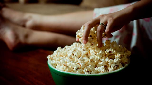 Eine Person greift mit der Hand in eine große grüne Schüssel voller Popcorn. Im Hintergrund sind unscharf die Beine der Person zu sehen, die offenbar auf einem Sofa sitzt. Die Szene vermittelt eine entspannte Atmosphäre, möglicherweise beim Filmschauen.