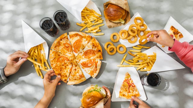 Eine Gruppe von Menschen teilt eine Mahlzeit auf einem Tisch. In der Mitte befindet sich eine Pizza mit Tomatenscheiben, umgeben von Pommes frites in Papiertüten, Zwiebelringen und einigen Gläsern Cola. Eine Person hält einen Zwiebelring, während eine andere ein Stück Pizza nimmt. Die Szene ist von Sonnenlicht durch Schatten von Blättern beleuchtet.