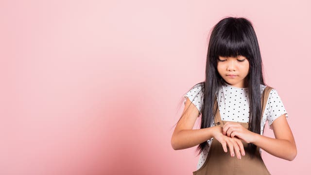 Ein junges Mädchen mit langen, dunklen Haaren steht vor einem rosa Hintergrund und kratzt sich am Arm. Sie trägt ein weißes, gepunktetes T-Shirt und einen braunen Overall.