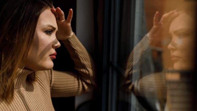 Eine Frau in einem beigefarbenen Rollkragenpullover steht nachdenklich an einem Fenster und schaut hinaus. Ihr Spiegelbild ist im Glas sichtbar. Die Szene ist in warmes Licht getaucht, was eine nachdenkliche und ruhige Atmosphäre schafft.