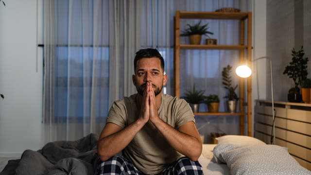 Ein Mann sitzt nachdenklich auf einem Bett in einem Schlafzimmer. Er trägt ein beiges T-Shirt und karierte Pyjamahosen. Im Hintergrund sind ein Regal mit Pflanzen und eine leuchtende Stehlampe zu sehen. Der Raum ist durch einen Vorhang abgedunkelt.