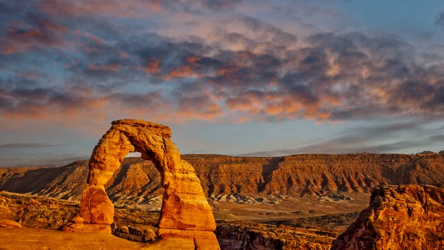 Eine beeindruckende Landschaftsaufnahme des Delicate Arch im Arches-Nationalpark, Utah, bei Sonnenuntergang. Der ikonische Sandsteinbogen steht im Vordergrund, während die untergehende Sonne die Felsen in warmen Orangetönen erstrahlen lässt. Der Himmel ist mit dramatischen Wolkenformationen bedeckt, die in sanften Rosa- und Blautönen leuchten. Im Hintergrund erstreckt sich eine weite, felsige Wüstenlandschaft.