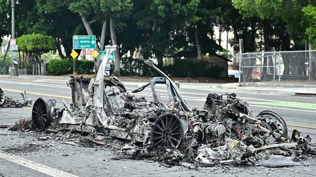 Ein ausgebranntes Autowrack steht auf einer Straße, umgeben von Trümmern. Im Hintergrund sind Straßenschilder zu sehen, die nach Norden und Süden weisen. Bäume und ein Zaun säumen die Straße. 
