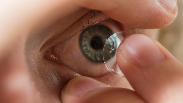 Eine Person setzt eine Kontaktlinse auf ein Auge. Die Nahaufnahme zeigt die Linse, die mit den Fingerspitzen gehalten wird, während das Auge weit geöffnet ist. Die Details der Iris sind deutlich sichtbar.