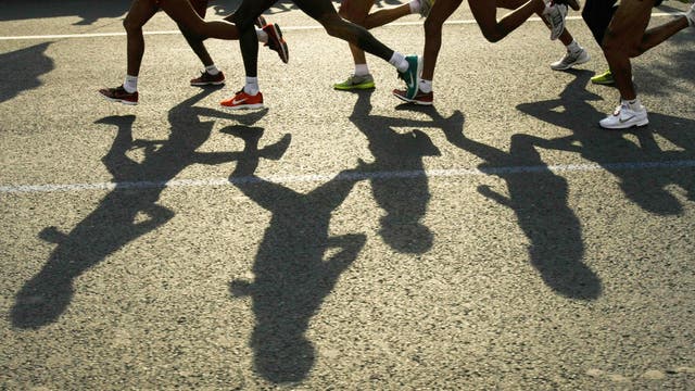 Mehrere Läufer in Bewegung auf einer Straße, ihre Schatten fallen deutlich auf den Asphalt. Die Szene zeigt dynamische Bewegung und Energie, während die Läufer in verschiedenen farbigen Sportschuhen laufen. Die Perspektive betont die Schatten der Läufer, die sich auf dem Boden abzeichnen.