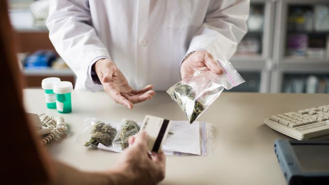 Eine Person in einem weißen Laborkittel übergibt einer anderen Person eine Plastiktüte mit getrockneten Pflanzen, möglicherweise medizinischem Cannabis. Auf dem Tisch liegen weitere Tüten mit ähnlichem Inhalt sowie zwei Pillenflaschen. Die Person im Vordergrund hält eine Kreditkarte. Im Hintergrund sind Regale mit unscharfen Gegenständen zu sehen.