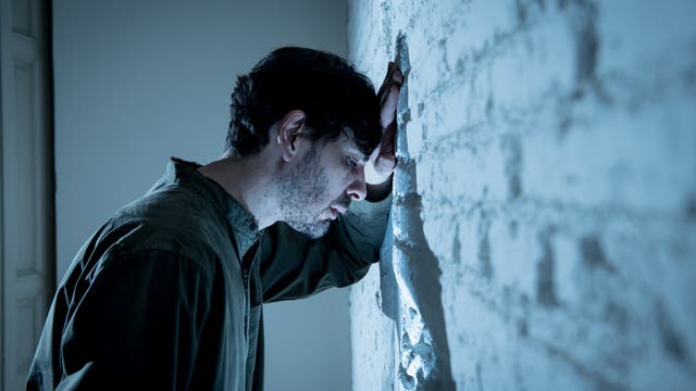 Ein Mann lehnt mit dem Kopf gegen eine weiße Backsteinwand, wirkt nachdenklich und bedrückt. Die Szene ist in kühlen Blautönen gehalten, was eine melancholische Stimmung vermittelt.