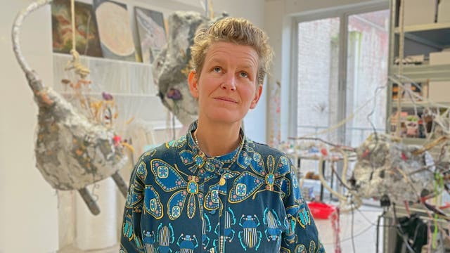 Eine Person steht in einem Atelier, umgeben von abstrakten Skulpturen und Kunstwerken. Sie trägt ein Hemd mit buntem, geometrischem Muster und blickt nachdenklich nach oben. Im Hintergrund sind verschiedene Kunstmaterialien und Werkzeuge zu sehen. Der Raum ist hell erleuchtet durch ein großes Fenster.