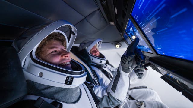Zwei Astronauten in weißen Raumanzügen sitzen in einem Raumschiffcockpit. Der vordere Astronaut lächelt, während der hintere Astronaut auf einen großen Bildschirm zeigt, der mit blauen Grafiken und Daten gefüllt ist. Die Szene vermittelt eine Atmosphäre von Technologie und Entdeckung.