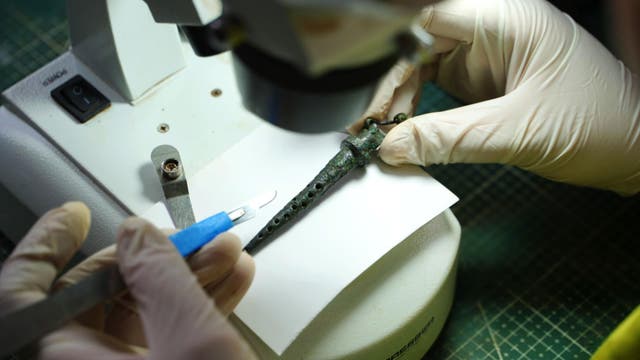 Eine Person mit weißen Handschuhen arbeitet an einem antiken Artefakt unter einem Mikroskop. Das Artefakt liegt auf einem weißen Blatt Papier, während die Person ein kleines Werkzeug in der Hand hält. Der Arbeitsplatz hat eine grüne, karierte Unterlage.